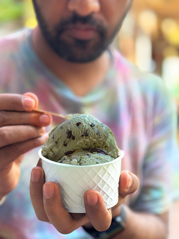 Stock photo showing a close-up view of street food dessert of scoops of mint choc chip ice cream served in a white, single-use, disposable, cardboard tub with wooden spoon.
