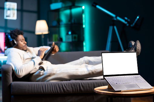 Man enjoying himself, searching new tv series to watch in apartment living room, focus on mockup laptop. Relaxed person wearing headphones, lying on couch next to isolated screen device