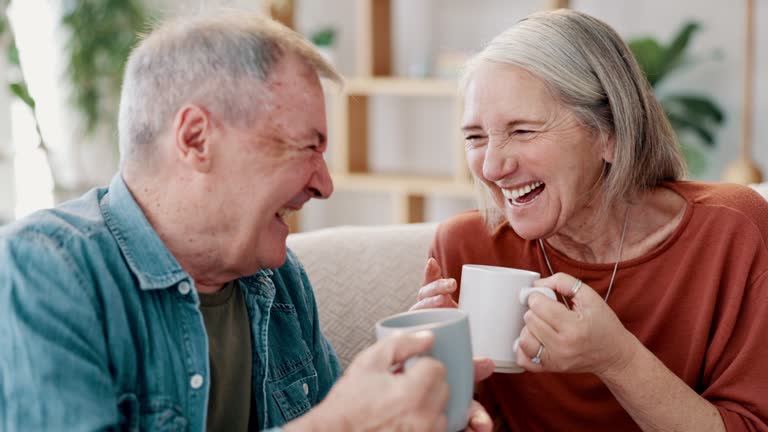 Senior couple, drinking coffee and love or laughing together at home, bonding and connection. Elderly people, forehead touch and tea for funny conversation on couch, happy and cheerful in living room