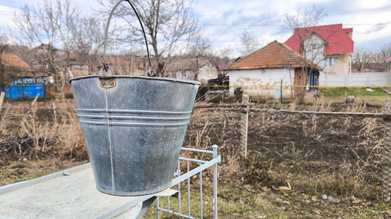Old metal bucket near the fountain on the background of an old house in the village.
