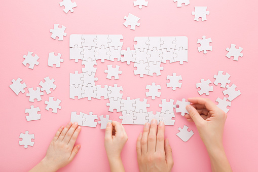 Mother and daughter hands playing and assembling white puzzle pieces on pink table background. Pastel color. Family activities. Spending time together. Closeup. Point of view shot. Top down view.