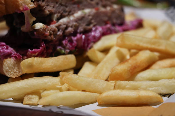 Stock photo showing close-up view of a meal of half a cheeseburger in a sesame seed bread bun served with a beef patty on bed of red cabbage coleslaw, a slice of melting processed cheese and crunchy French fry chips.