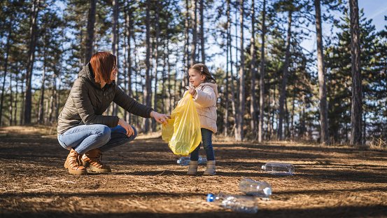 mother and daughter small caucasian girl toddler and woman collect plastic bottle waste garbage from the forest nature park put it in the bag