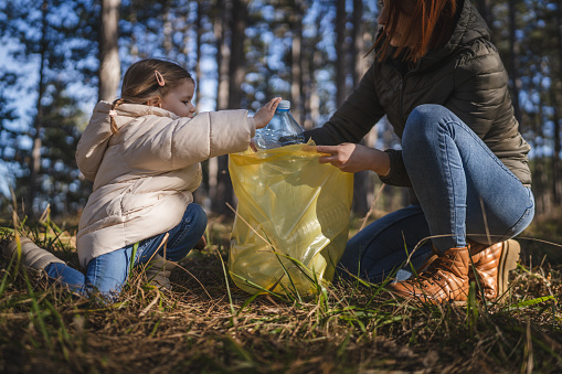 mother and daughter small caucasian girl toddler and woman collect plastic bottle waste garbage from the forest nature park put it in the bag