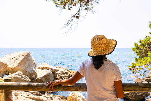 Unknown woman leaning on a wooden railing and wearing a hat enjoying a sensational view towards the sea surrounded by pine trees on a summer day on the Spanish Costa Brava