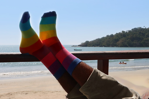 Stock photo showing close-up view of man's feet crossed at ankles on wooden railing, wearing blue, turquoise, green, yellow, orange, red, pink and purple striped socks.