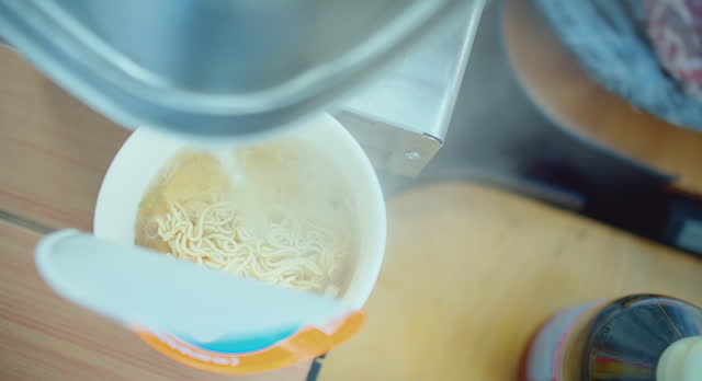 Overhead view of instant noodles being prepared with steaming hot water poured from an electric kettle, Hot water pouring into a cup of instant noodles, a quick meal preparation