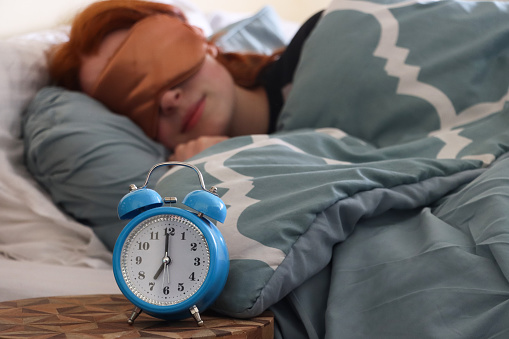 Stock photo showing close-up view of a blue metal case retro alarm clock with double bell on night stand. An attractive, redheaded woman can be seen lying in the double bed, next to the beside table, sleeping with an eye mask.