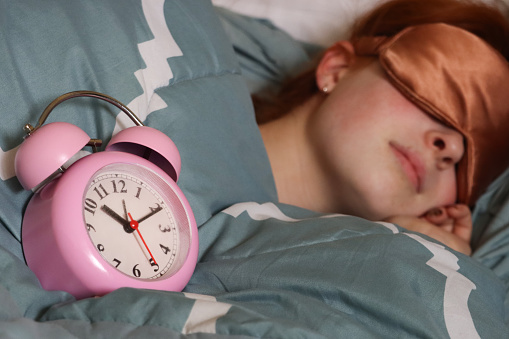 Stock photo showing close-up view of a pink metal case retro alarm clock with double bell on duvet. An attractive, redheaded woman can be seen lying in the double bed, sleeping with an eye mask.