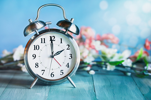Set your clocks forward with this clock and flowers over a rusitc teal wooden table. Daylight saving time concept. Selective focus with blurred background.