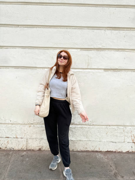 Image of attractive, redheaded woman wearing sunglasses, fluffy jacket, jogging trousers, crop top, tote bag on shoulder, smiling looking at camera, white wall background, focus on foreground, copy space Stock photo showing close-up view of red haired woman standing outdoors before a white wall, smiling at camera. corduroy jacket stock pictures, royalty-free photos & images