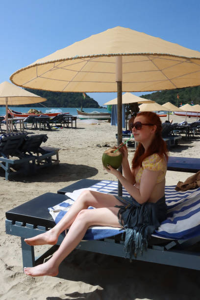 晴れた日にサングラスをかけた長い髪の魅力的な赤毛の女性の画像、ビーチパラソルの日傘の下に座り、ストローを飲むココナッツ、砂浜の背景、横顔図、前景に焦点を当てる - outdoor chair beach chair sarong ストックフォトと画像