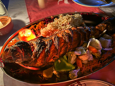 Stock photo showing close-up, elevated view of an oval, metal platter of tandoori marinated, grilled red snapper fish served with rice and vegetables and a tomato candle.
