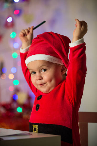 Charming little boy in Santa costume sitting with raised arms at table while holding pencil and looking at camera in decorated room