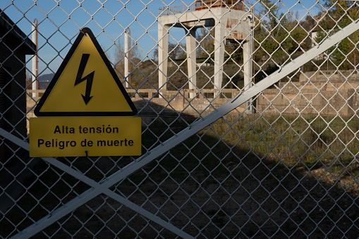 At the entrance to a fenced-off area, a small electrical substation has been posted with a yellow warning sign, high voltage danger to life. Danger sign.