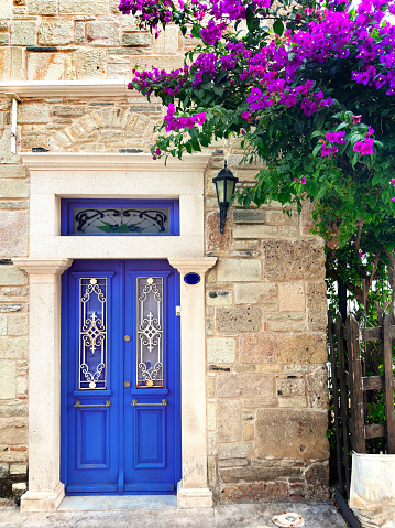 Vintage white bike with flowers in a basket against the blured background of a white mediterranean house with a blue door and window and flowering tree on the wall.