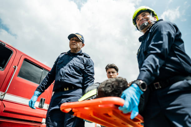 Two first responders giving medical attention to a teenage boy lying on a stretcher outdoors. The paramedic, a mature man in his 50s, and emergency medical technician, an Hispanic woman in her 40s, work together to care for the injured or ill patient.