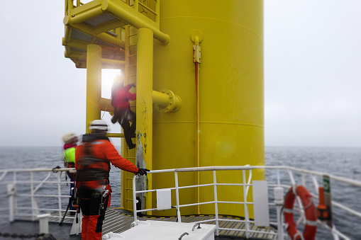Industrial climber,  rope access technician climbing on ladder on gigantic offshore wind turbine with sun behind him from passenger ship deck and another technicians checking him. Borkum riff Grund, germany.