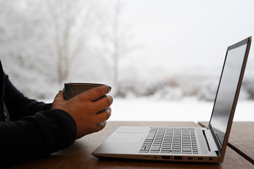 Person holding a coffee mug, taking a break from working remote on laptop. Working from home on a winter day with snow outside. Photo taken in Sweden.