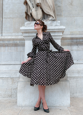 Full length shot of a young brunette in a dotted dress on high heels leaning against wall  in Paris.Establishing  shot.