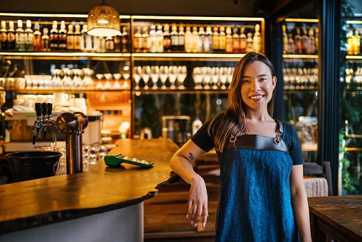 Portrait of a smiling young female bartender wearing an apron leaning against a checkout counter in a trendy bar
