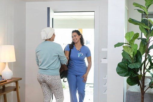 An unrecognizable female cancer patient welcomes the young adult female hospice nurse at the front door of her home.