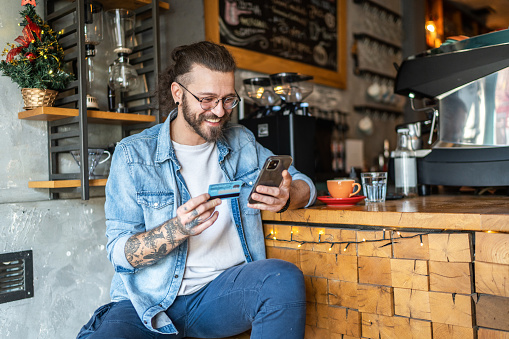 Smiling young man sitting at coffee shop and shopping online