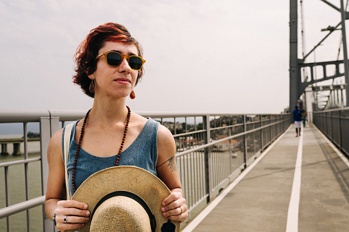 Young woman wearing sunglasses and holding a sun hat looking at the scenic view from Hercilio Luz bridge, in Florianopolis, Brazil