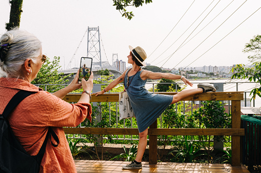 Smiling mature woman taking photos of her adult daughter posing on an observation point with a scenic bridge in the background