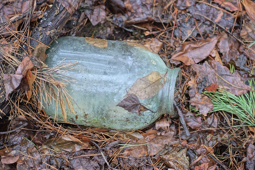 trash from one big gray dirty glass jar lies in brown wet fallen leaves in nature