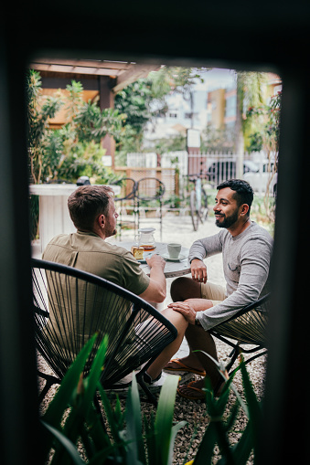 Smiling young gay couple talking together at a table in a coffee shop patio in the early morning