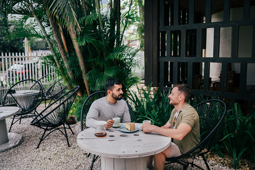 Smiling young gay couple talking together at a table in a coffee shop patio in the early morning