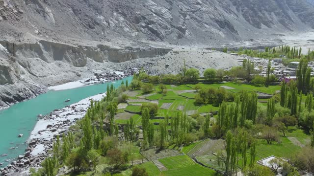 Drone flying over terraced fields, tall trees and towards green water of a glacial river flowing besides mountains.