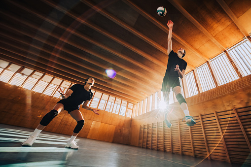 Bottom view of a teenage female professional volleyball players in action hitting a ball on indoor court. Lens flare. Low angle view of a multicultural professional sportswomen hitting a volleyball.