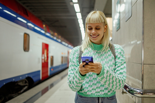 Happy passenger is standing at metro station and smiling at the phone while typing message on it. A hipster girl arrived to her destination so she is typing a message to her friend. Near girl is train