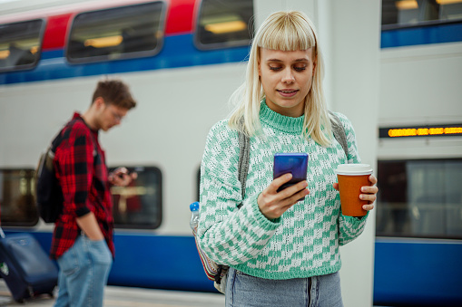 An urban girl is standing at metro station with coffee to go in her hands and typing on her phone. A young passenger is looking at her phone at train station. In a blurry background is a metro train.
