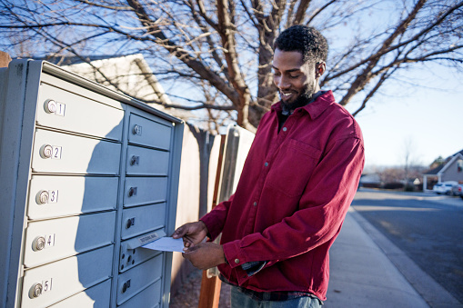 Young Cheerful African-American Man In His Twenties Voting By Mail with Ballot at a Neighborhood Mail Box