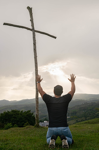 Man on his knees raising his hands and praying to god in the mountains. Wooden cross with sacred sky background