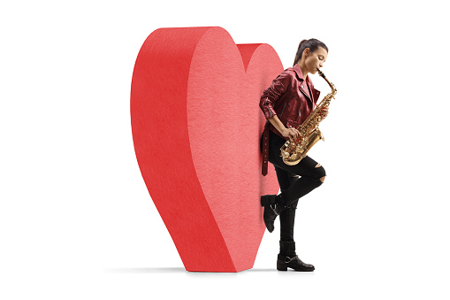 Young woman playing saxophone and leaning on a red heart, love and romance concept isolated on white background
