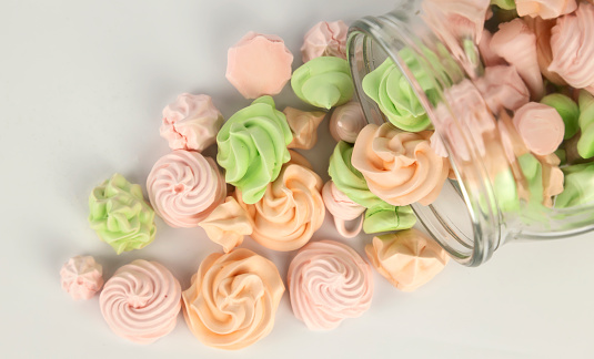 Many colorful meringues pouring out of a glass jar on a white background, close-up, top view.