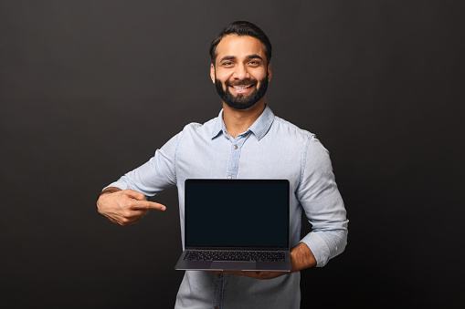 Happy Indian man holding laptop with empty screen and points finger at it, hispanic male employee presenting new computer app, promotion and advertise concept