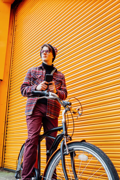 istock Young hipster man using mobile phone while sitting on cruiser bicycle near orange wall. Student using 5g network,rests after riding. Eco friendly transport for carbon footprint. Sustainable lifestyle 1991376466