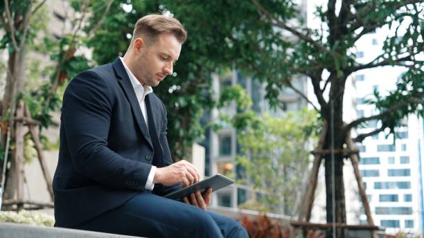 istock Businessman sitting at park while working on tablet by reading report. Urbane 1991538174