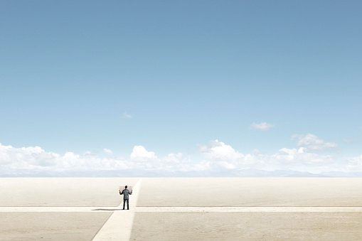 A man stands at a crossroad while holding and looking at a road map as he considers what path will take him to his destination.