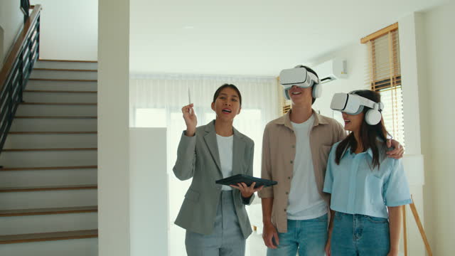 Asian female real estate agent explaining and showing by using VR and tablet in new house to tenants for rent or buy. Landlord selling real estate to happiness couple customer.