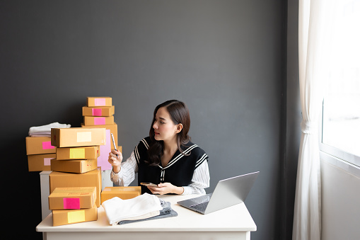 Asian female online seller Count products smartly using a pen and holding a smartphone. It is the same amount as the customer confirmed. Big business in the export industry Working in the office