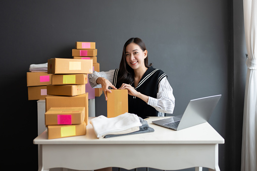 Asian female online seller opens big business in parcel and order export industry Unpacking merchandise to check for completeness for customers. There is a laptop and clothes on the table.