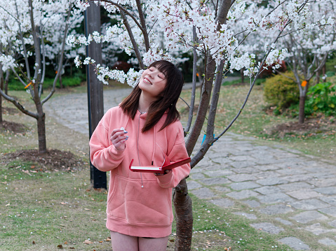 Beautiful young Chinese woman smiling with eyes closed under blooming cherry tree, wearing pink loose oversized top and holding red notebook. Emotions, people, beauty, youth and lifestyle portrait.