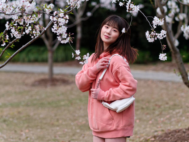 istock Beautiful young Chinese woman smiling under blooming cherry tree, wearing pink loose oversized top with small white shoulder bag. Candid moment. Emotions, people, beauty, youth and lifestyle portrait. 1991781149