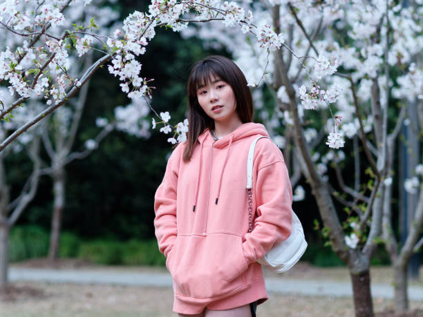 istock Beautiful young Chinese woman posing under blooming cherry tree, wearing pink loose oversized top with small white shoulder bag. Candid moment. Emotions, people, beauty, youth and lifestyle portrait. 1991799054